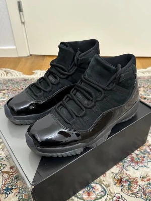 Svarta Air Jordan 11 sneakers - Säljer ett par svarta Air Jordan 11 sneakers med blankt lackat läder och mockadetaljer. Skorna har högt skaft, tjocka svarta skosnören och en robust sula. Perfekta för dig som gillar klassisk basketstil och vill sticka ut med en ikonisk modell.