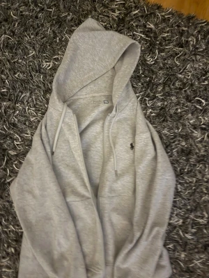 Grå hoodie från Polo Ralph Lauren - Säljer en grå hoodie från Polo Ralph Lauren med dragkedja och huva. Klassisk design med logotyp broderad på bröstet och snören i huvan. Materialet är mjukt och bekvämt, perfekt för chill dagar. Passar dig som gillar stilrena och tidlösa plagg. Köpt från butiken för 1750kr mitt pris 800kr, pris kan diskuteras vid snabbaffär!