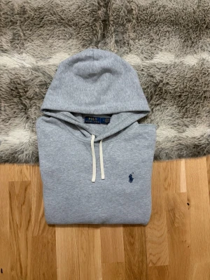 Grå hoodie från Polo Ralph Lauren - Snygg grå hoodie från Polo Ralph Lauren med klassisk broderad logga på bröstet. Hoodien har vit dragsko, känguruficka och ribbade muddar. Tillverkad i mjuk bomullsblandning som är skön att ha på sig. Perfekt för en avslappnad och stilren look.