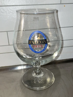 Falcon Beer ölglas med logga - Säljer ett stort ölglas från Falcon Beer med tydlig logga i blått, svart och guld samt texten 'Bryggt på rent källvatten'. Glaset har rundad form och stabil fot, perfekt för att njuta av en kall öl.