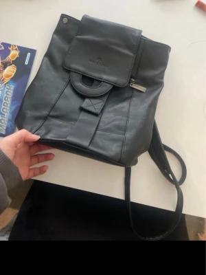 Svart ryggsäck  - Stilren svart ryggsäck i slätt konstläder med lock och dragkedja. Väskan har en enkel design med justerbara axelband och en dekorativ rem på framsidan. Perfekt för att bära dina grejer till skolan eller stan.