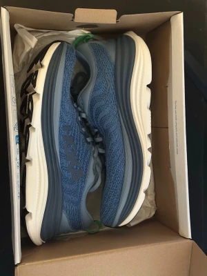 Blå Hoka sneakers med mesh - Snygga blå sneakers från Hoka med stickad mesh-överdel och vita sulor. Skorna har gröna detaljer på hälen och klassiska snörningar. Perfekt för dig som gillar sportig stil och komfort. Hoka-loggan syns tydligt på sidan.