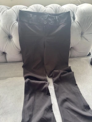 Svarta kostymbyxor med dubbelknäppning - Stilrena svarta kostymbyxor med dubbelknäppning framtill och raka ben. Byxorna har två fickor med knapp på baksidan och är tillverkade i ett slätt material som ger en snygg och klassisk look. Perfekta för dig som vill ha en clean och modern stil.