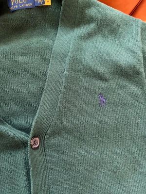Polo Ralph Lauren Cardigan - Tja! Säljer nu en stilren mörkgrön cardigan från Polo Ralph Lauren i otroligt fint skick,  fem knappar framtill och i frägen mörkgrön. Nypris ca 3000 kr, säljer för knappt halva! Två fickor framtill och ribbade muddar vid ärmslut och nederkant. Perfekt för lager på lager och enkel att matcha. Storlek M. Pris går alltid att diskuteras✅. 