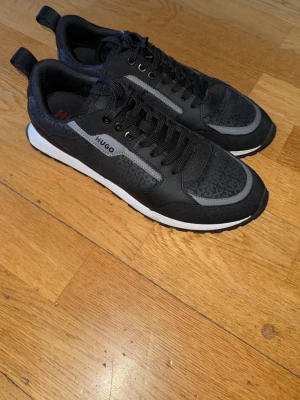 Svarta sneakers från Hugo - Snygga svarta sneakers från Hugo med diskret logomönster på sidan och grå detalj längs snörningen. Skorna har svart ovandel i textil och syntet, vita sulor och svarta snören. Perfekta för en trendig och sportig look.