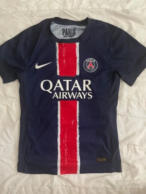 PSG matchtröja blå Nike - Snygg blå PSG matchtröja från Nike med röd och vit rand i mitten, klubbmärke och Qatar Airways-tryck på bröstet. Tillverkad i lätt och ventilerande material med Dri-FIT ADV-teknologi. Perfekt för fotboll eller träning. Pris kan diskutera