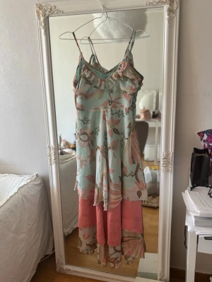 Blommig långklänning med volang - Långklänning med tunna justerbara axelband och volangkant upptill. Klänningen har ett blommigt mönster i ljusblått, rosa och grönt samt en bred rosa volang nedtill. Tillverkad i ett tunt, luftigt material som känns som polyester eller bomull.