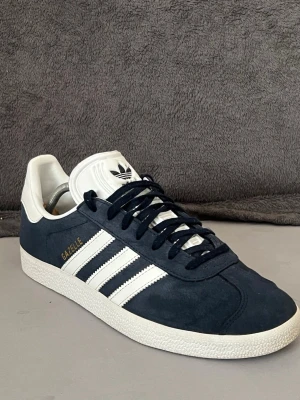 Adidas Gazelle navy sneakers - Snygga Adidas Gazelle sneakers i mörkblå mocka med vita detaljer och klassiska tre ränder på sidan. Skon har vit sula och vit häl, samt guldfärgad Gazelle-text. Perfekt för dig som gillar retrostil och ikoniska modeller.