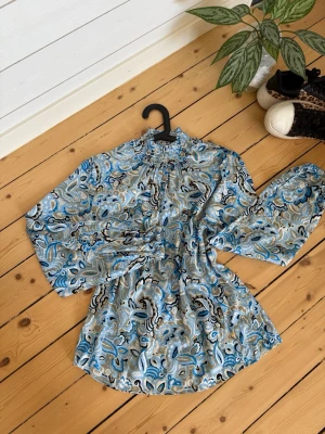 Blå paisleymönstrad blus - Snygg blus med blått, grått och svart paisleymönster. Blusen har hög krage med rynkade detaljer och långa ärmar med resår vid handlederna. Materialet är lätt och luftigt, perfekt för att skapa en cool look.