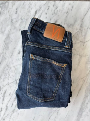 Nudie Jeans grimtim - Säljer ett par mörkblå jeans från Nudie Jeans med klassisk femficksdesign och kontrastsömmar. Jeansen har en slim fit och är tillverkade i bomull med läderpatch bak. Perfekt för dig som gillar stilrena och tidlösa jeans.