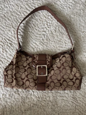 Väska oanvänd - Cool axelväska i beige och brunt med klassiskt monogrammönster och ett stort silverfärgat spänne på framsidan. Väskan har läderdetaljer och en bred axelrem som gör den enkel att bära. Perfekt för dig som gillar stilrena accessoarer.