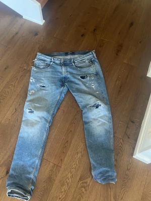 Blå slitna jeans med hål - Säljer ett par blå jeans med slitningar och hål på flera ställen. Jeansen har en klassisk femficksmodell och raka ben. Perfekta för en avslappnad och trendig look. Materialet är denim i bomull och färgen är ljusblå med tvättade detaljer.
