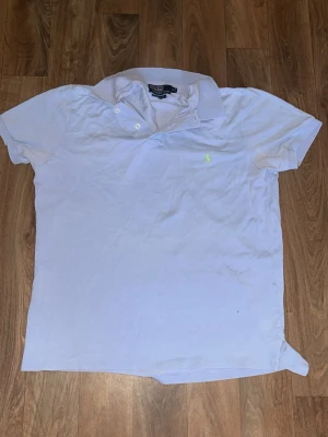 Blå pikétröja från Ralph Lauren - Klassisk blå pikétröja från Ralph Lauren med korta ärmar och krage. Tröjan har två knappar vid halsen och den ikoniska broderade loggan i ljusgrönt på bröstet. Perfekt till jeans eller shorts för en clean och stilren look.