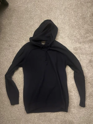 Mörkblå hoodie från Oscar Jacobson - Snygg mörkblå hoodie från Oscar Jacobson med klassisk huva och dragsko. Tillverkad i mjukt material som känns skönt mot huden. Perfekt för dig som gillar stilrena och enkla plagg med en modern touch. Långa ärmar och smal passform.