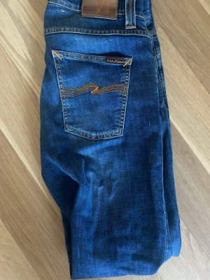 Mörkblå jeans från Nudie Jeans - Snygga mörkblå jeans från Nudie Jeans modell: Lean Dean med klassisk femficksdesign och orangea sömmar på bakfickorna. Jeansen är tillverkade i denim av bomull. Perfekta för en avslappnad och stilren look. Jeansen har ett hål som man ser på bilden fast det är enkelt att fixa, Jag kan inte sy 🫠storlek 30/32. Skriv till mig om ni har några frågor eller vill har bättre bilder😁