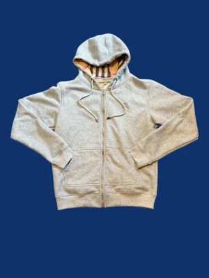 Grå zip-hoodie från Burberry - Säljer en grå hoodie från Burberry med dragkedja och klassiskt rutmönster i huvan. Tröjan har två fickor fram, ribbade muddar och snören i huvan. Perfekt för dig som gillar stilrena och exklusiva plagg. Det står M, men passar S/xs så tänk på de om ni köper den.