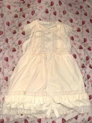 Söt vit kortklänning med spetsdetaljer - Supersöt vit kortklänning med spets och volanger, knappar framtill och korta ärmar. Klänningen har romantisk stil och är perfekt nu inför våren och sommaren🩷 Använd ca 2 gånger.