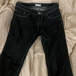 Svarta bootcut jeans från Gina Tricot - Säljer ett par svarta bootcut jeans från Gina Tricot med vita kontrastsömmar och klassisk femficksdesign. Jeansen har låg midja och vida ben, perfekt för dig som gillar en retrostil. Materialet är denim och de har knapp- och dragkedjeförslutning. Storlek 38, använda en gång, säljer pga för små. Köpte för ca 400kr