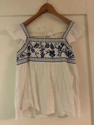Vit blus med blå broderier - Söt vit blus med blå broderade blommor och rutmönster upptill. Blusen har korta ärmar, fyrkantig ringning och knytband i ryggen. Tillverkad i luftig bomull, perfekt för varma dagar. Ger en romantisk och bohemisk vibe.