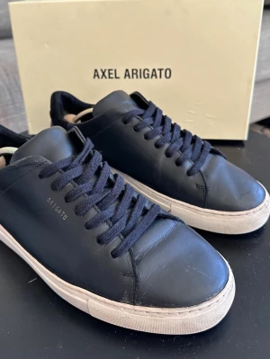 Svarta Axel Arigato i läder - Snygga svarta sneakers från Axel Arigato i äkta läder med klassisk vit sula och svarta skosnören. Minimalistisk design med diskret logotyp på sidan. Perfekta för dig som gillar stilrena och trendiga skor. Skorna är i bra skick men finns täcken på användning som bilderna visar, som man ser är de enda lila defekter på sulan, men ingen som syns när man har de på.