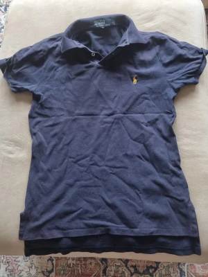 Marinblå pikétröja från Ralph Lauren - Klassisk marinblå pikétröja från Ralph Lauren med korta ärmar och den ikoniska broderade loggan på bröstet. Tillverkad i mjuk bomull och har en snygg krage med knappslå. Perfekt till jeans eller kjol för en clean look.