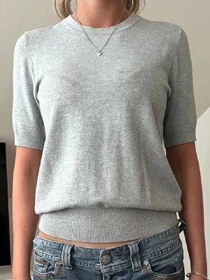 Ljusgrå stickad t-shirt  från Zara - En stilren ljusgrå topp från Zara med rund halsringning och korta ärmar. Toppen har en ribbad nederkant och är tillverkad i ett mjukt material som känns skönt mot huden. Perfekt att matcha med jeans eller kjol.