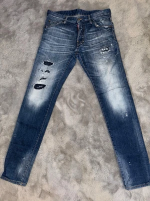 Blå slitna jeans från Dsquared2 - Säljer ett par blå jeans från Dsquared2 med slitningar och hål på framsidan. Jeansen har klassisk femficksmodell, låg midja och snygga slitningar för en trendig look. Materialet är denim i bomull med stretch för extra komfort.