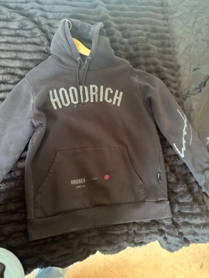 Svart hoodie från Hoodrich - Svart hoodie från Hoodrich med stor logga framtill och tryck på ärm och rygg. Klassisk känguruficka och dragsko i huvan. Mjuk bomullsblandning, perfekt för en streetwear-look.