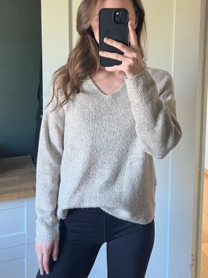 Beige stickad v-ringad tröja - Mysig beige stickad tröja med v-ringning och lång ärm. Tröjan har en avslappnad passform och ribbad struktur som ger en skön känsla. Perfekt att matcha med jeans eller tights för en enkel och snygg look.