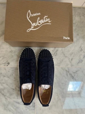 Christian Louboutin svarta sneakers - Snygga svarta sneakers från Christian Louboutin med klassiska röda sulor och coola nitar på tån. Skorna har en låg profil, snörning och är tillverkade i mocka med läderdetaljer. Perfekt för dig som vill sticka ut med exklusiva och trendiga skor.