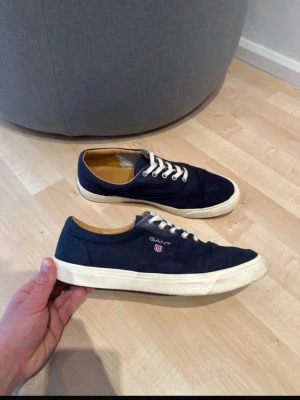 Marinblå sneakers från Gant Strl 42 - Snygga marinblå sneakers från Gant med vit sula och vita skosnören. Skorna har en klassisk låg modell och är tillverkade i canvas med detaljer i beige på insidan. Perfekta för en stilren och avslappnad look. Använda i bra skick, lättare sliten i sul hälen se bild, storlek 42, pris kan diskuteras 