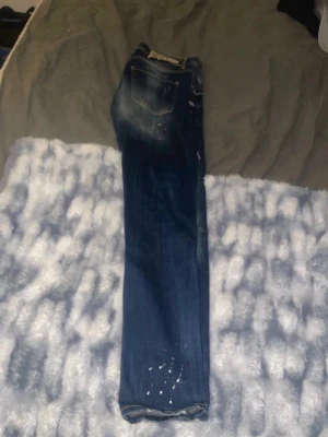 Mörkblå slitna jeans med hål - Säljer ett par mörkblå jeans med slitningar och hål på båda benen. Jeansen har en tvättad look med ljusare partier och vita färgstänk för en trendig, avslappnad stil. Perfekta för dig som gillar streetwear och en edgy vibe. Osäker på storlek om ni vill ha mått är de bara att fråga