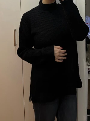 Svart stickad polotröja - Säljer en svart stickad polotröja med lång ärm och avslappnad passform. Tröjan har hög krage och är ribbstickad vid ärmslut och nederkant. Perfekt för lager-på-lager och enkel att matcha med jeans eller kjol.