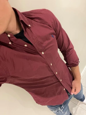 Vinröd skjorta från Ralph Lauren - Vinröd skjorta med button-down krage från Ralph Lauren. Långärmad modell med vita knappar och blå broderad logga på bröstet. Skjortan är i bomull och har en klassisk, stilren look som passar till jeans eller chinos.