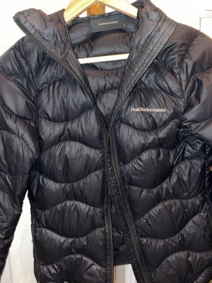 Svart pufferjacka Peak Performance XS - Snygg svart pufferjacka från Peak Performance med vågformat quiltat mönster och dragkedja framtill. Jackan har hög krage och diskret logga på bröstet. Tillverkad i lätt, glansigt material som håller värmen utan att kännas tung.