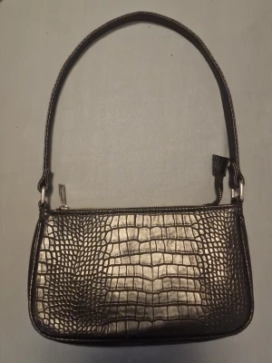 Svart krokopräglad axelväska - Stilren svart axelväska i syntetmaterial med krokodilpräglad yta och silverfärgad dragkedja. Väskan har en rektangulär form och ett kort axelband, perfekt för att lyfta din outfit med en trendig touch.