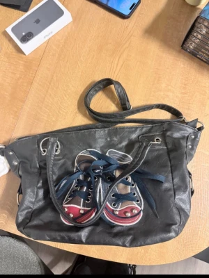 Svart axelväska med sneakersmotiv - Cool svart axelväska i konstläder med unikt broderat sneakersmotiv framtill och blå skosnören som detalj. Väskan har justerbar axelrem, dragkedja upptill och rymligt innerfack. Perfekt för dig som gillar streetstyle och vill sticka ut.