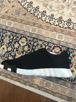 Svarta H skor - Stilrena svarta sneakers med mesh-överdel och detaljer i mocka. Vit sula med svart logga. Skorna har en chunky, räfflad vit sula och en svart detalj på hälen. Lågt skaft och modern design som passar till streetwear.