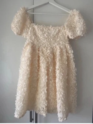 Krämvit puffärm klänning med 3D-blommor - Supersöt kort klänning i krämvit ton med puffärmar och massor av 3D-blommor över hela tyget. Klänningen har en luftig och voluminös form, perfekt för dig som vill sticka ut. Ingen synlig dragkedja eller knappar, stretchig rygg för extra komfort.