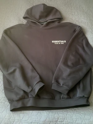 Svart Essentials hoodie Fear of God - Svart hoodie från Fear of God Essentials med vit logotyp på bröstet och ryggen. Klassisk passform med ribbade muddar och huva. Materialet är mjukt och bekvämt, perfekt för en avslappnad streetwear-look.