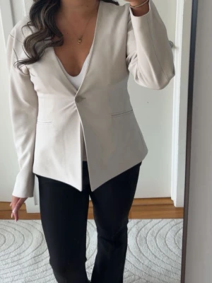 Beige kavaj med enkel knäppning - Stilren beige kavaj med en knapp framtill och figurnära passform. Kavajen har långa ärmar, diskreta fickor och en djup v-ringning. Perfekt för en trendig och clean look. Materialet ser ut att vara mjukt och lätt, vilket ger en snygg siluett.