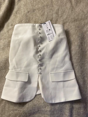 Vit kortkjol med knappar från Zara - Stilren vit kortkjol från Zara med dekorativa klädda knappar framtill och två fickdetaljer. Kjolen har en liten slits framtill och är gjord i ett lätt material som känns fräscht och somrigt.