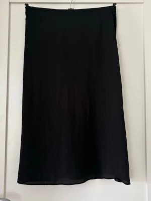 Svart långkjol i satinlook - En stilren svart långkjol med lätt glansig satinlook och lätt klockad passform. Kjolen har en enkel design utan synliga detaljer och faller snyggt längs benen. Perfekt för dig som gillar minimalistisk stil. Längd 77 cm.