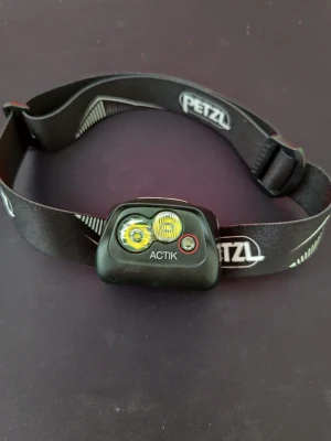 Petzl Actik pannlampa svart - Säljer en svart Petzl Actik pannlampa med justerbart elastiskt band och två LED-lampor. Lampan har en tydlig knapp för att slå på och av ljuset och är perfekt för utomhusaktiviteter som vandring eller löpning. Märket är tryckt på bandet.