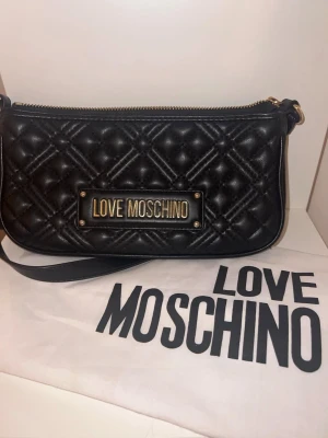  Moschino väska  - Snygg svart  handväska från Love Moschino med guldfärgade detaljer och logotyp framtill. Väskan har en axelrem och dragkedja upptill. Tillverkad med elegant mönster som ger en lyxig och premium känsla. Den är i mycket gott skick och har inga fläckar varken innuti eller utanpå väskan! 