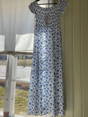 Vit långklänning med blå blommor - Oanvänd. Superfin långklänning i vitt med blått blommönster och smockad överdel. Klänningen har korta ärmar och en luftig, rak passform. Perfekt för varma dagar och har en romantisk känsla. Tillverkad i mjuk bomull som känns skön mot huden.