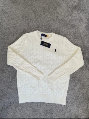 Vit kabelstickad tröja från Polo Ralph Lauren - Säljer en vit kabelstickad tröja från Polo Ralph Lauren med klassiskt svart broderat logotyp på bröstet. Tröjan har rund halsringning och ribbade muddar vid ärmslut och nederkant. Perfekt för dig som gillar stilrena och tidlösa plagg.