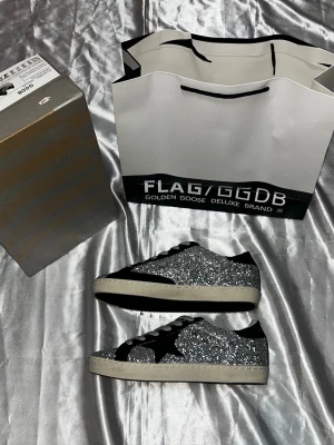 Glittriga sneakers från Golden Goose - Säljer ett par riktigt snygga sneakers från Golden Goose med silverglittrande ovandel, svart stjärndetalj på sidan och svarta samt vita detaljer. Skorna har vita sulor och grå skosnören. Perfekta för dig som vill sticka ut med din stil. 25cm 