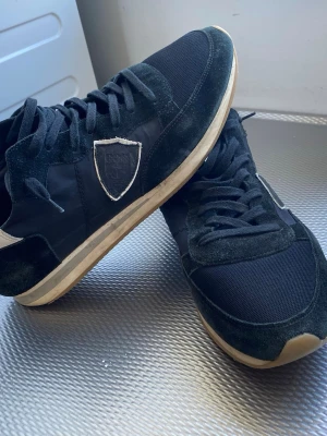 Svarta sneakers från Philippe Model - Det är lite slitna under skorna som ni ser på sista bilden annars är de i bra skick Snygga svarta sneakers från Philippe Model med klassisk låg profil och vit kant på hälen. Skorna har en kombination av mocka och textil, samt en vit emblem på sidan. Yttersulan är beige och ger en cool kontrast till den svarta ovandelen.