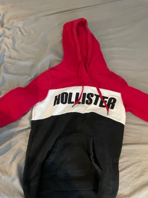 Hollister hoodie i rött, vitt och svart - Snygg hoodie från Hollister med färgblock i rött, vitt och svart. Stor logotyp på bröstet och ärmen, huva med dragsko och klassisk magficka. Perfekt för dig som gillar streetstyle och vill ha något bekvämt och trendigt. Materialet är mjukt och känns skönt mot huden.